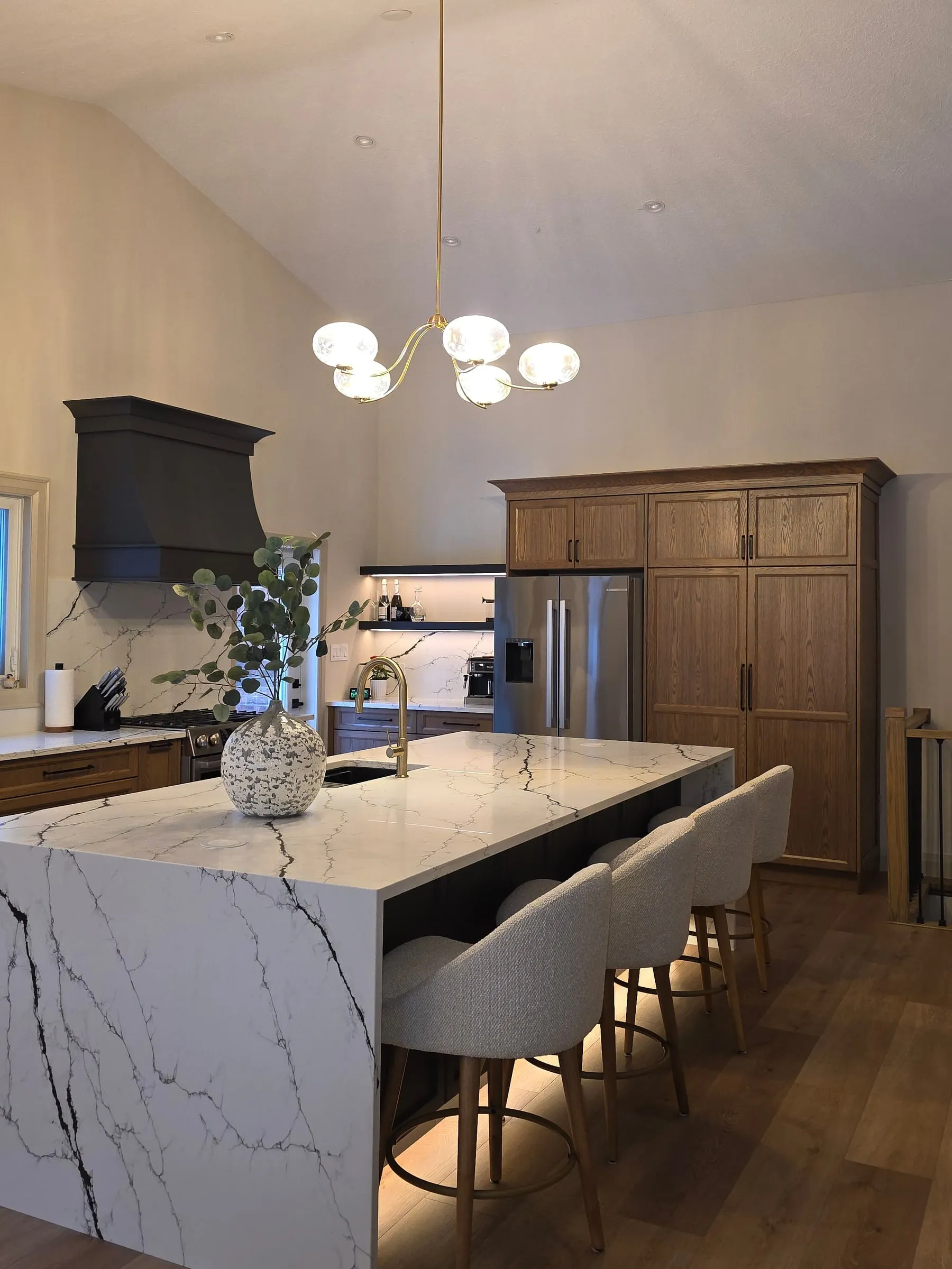 A kitchen island with a veined white waterfall counter, grey stools, a globe chandelier and wood cabinets behind